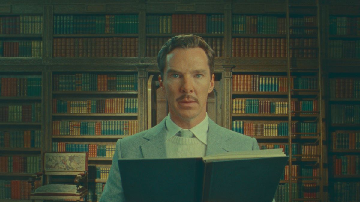 Benedict Cumberbatch, standing in a room lined ceiling to floor with books, stares directly into the camera while holding up another book in Wes Anderson’s Netflix film The Wonderful Story of Henry Sugar
