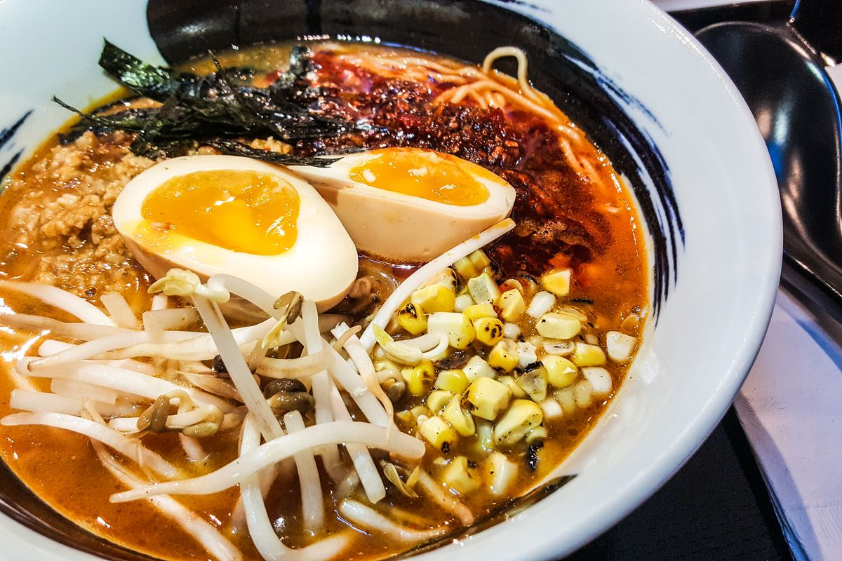 A white bowl with a thick, ink-like swoosh of black around the rim is filled with spicy miso ramen, including corn, bean sprouts, and egg, and other ingredients.