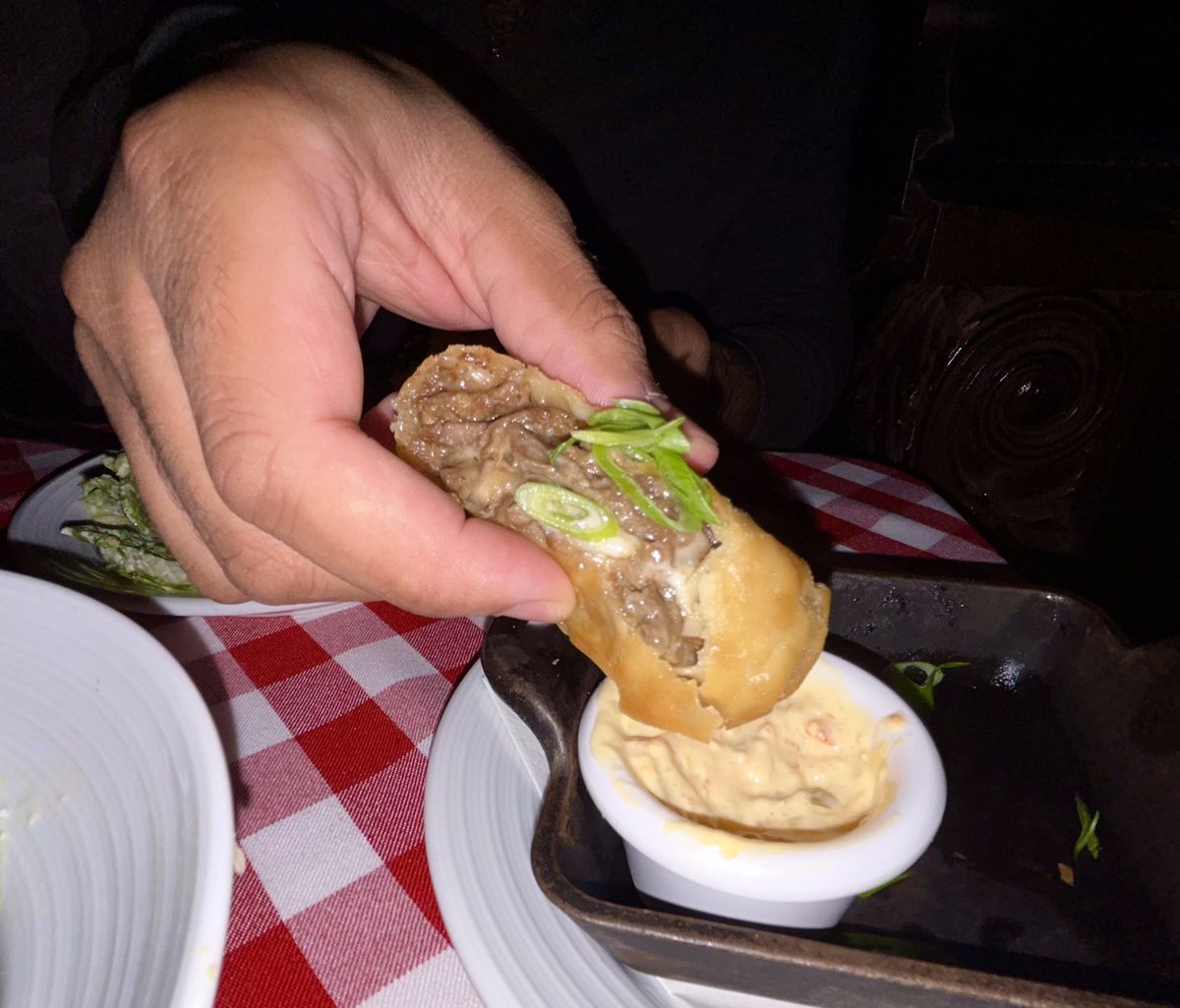 A hand dunks an egg roll filled with gray beef and American cheese into a pale sauce.