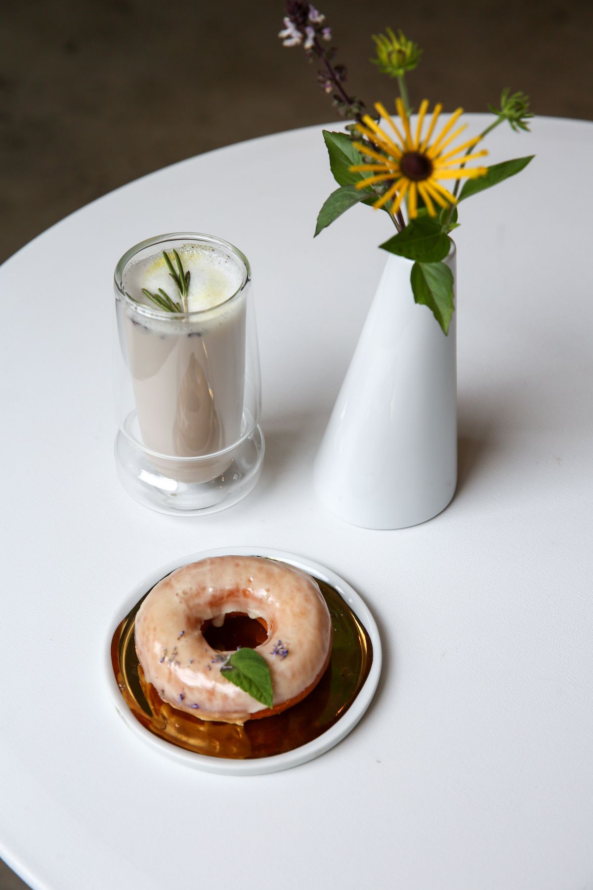 An image with a flower vase with a yellow flower, a white drink, and a white glazed doughnut.