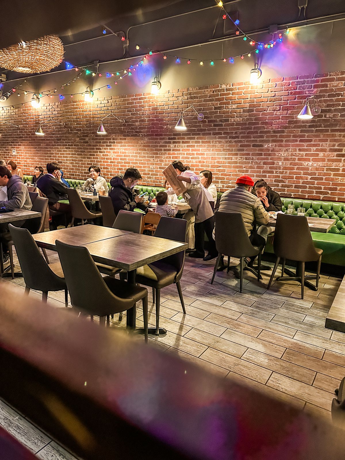 The interior of a busy sushi restaurant. Diners sit at tables against a brick wall with a string of holiday lights above.