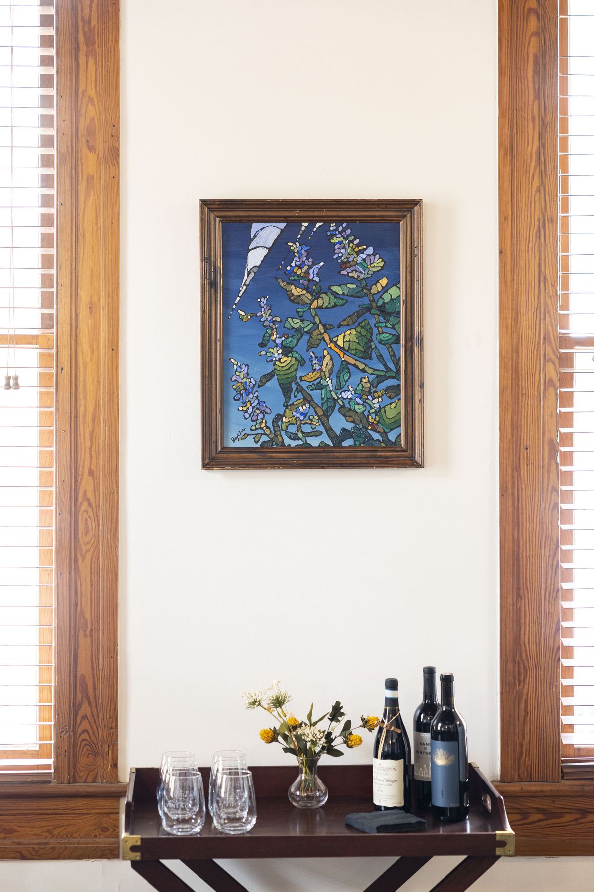 A wall with a painting and a table of stemless wine glasses and wine bottles.