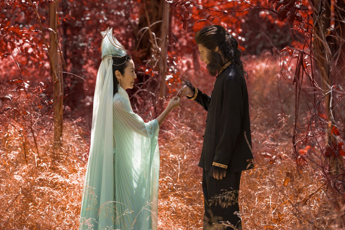 Michelle Yeoh and Jim Liu as Guanyin and Wei-Chen, son of the Monkey King, stand in a red forest as she hands him the Monkey King’s shrunken staff in American Born Chinese. 