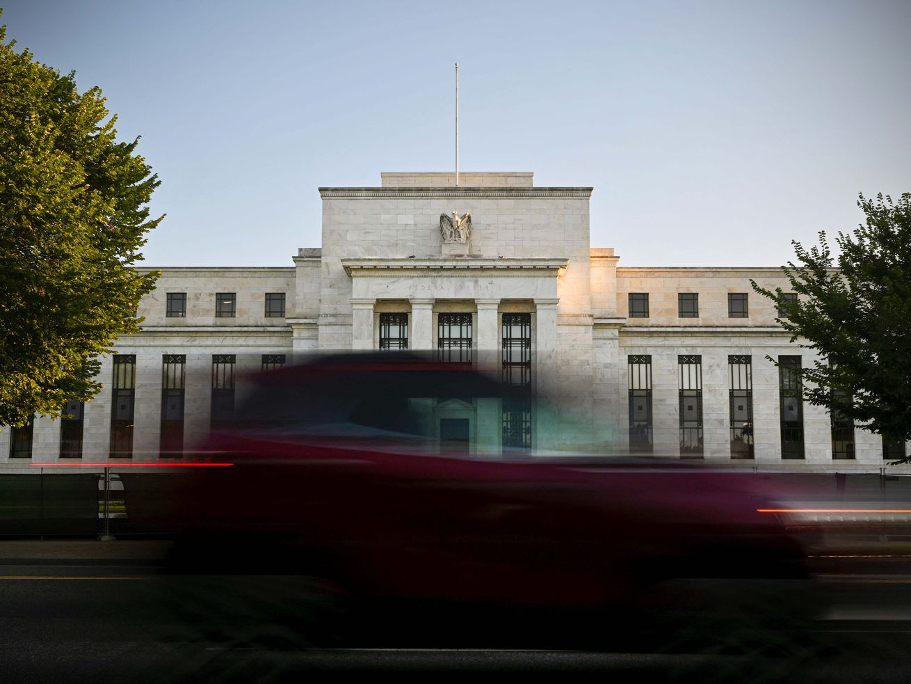 A time-lapse photo shows the Federal Reserve building, a white stone-fronted building with tall columns and vertical windows, as a blurred stream of traffic passes in front of it and the sunlight on the surrounding trees moves slowly.