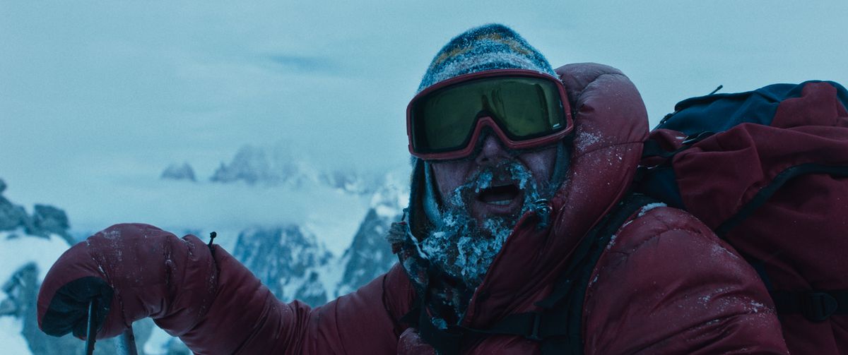A man in red climbing gear is covered with snow, especially his beard.  He wears a red backpack and looks above the camera, with the snowy mountains behind him.