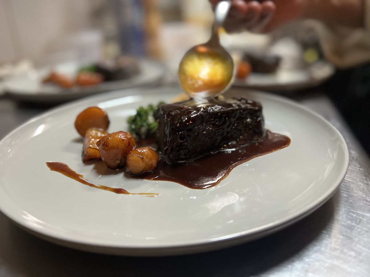 A spoon drizzles sauce on a short rib sitting on a white plate with orange vegetables arranged alongside.
