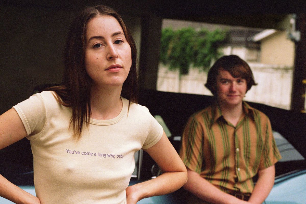 A young woman stands in the foreground wearing a T-shirt that reads “You’ve Come a Long Way, Baby.” In the background a teenage boy watches her.