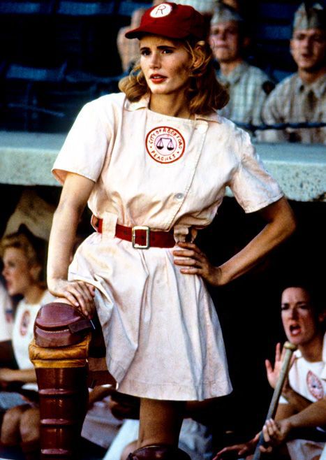 A blond woman in a peachy baseball uniform and catcher pads stands in front of a shelter.