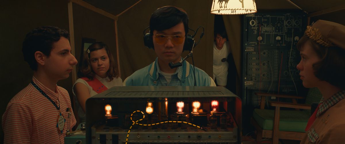 In a very dim room, a group of young people (Jake Ryan, Grace Edwards, Ethan Josh Lee, Aristou Meehan, and Sophia Lillis) stand around an old-timey radio covered with lights and dials in Wes Anderson's Asteroid City