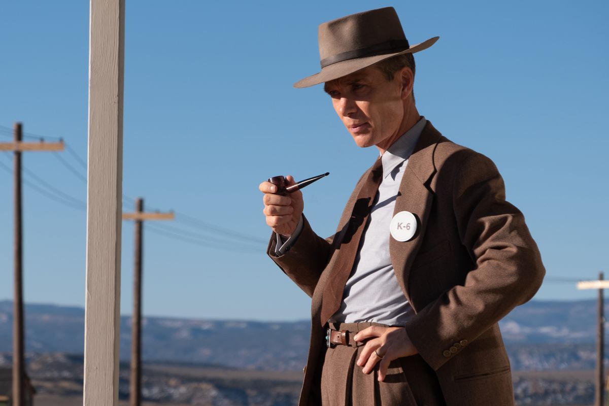 J. Robert Oppenheimer (Cillian Murphy), in brown suit and hat, holds a pipe and stands in a desert near a row of telephone poles in Christopher Nolan’s Oppenheimer