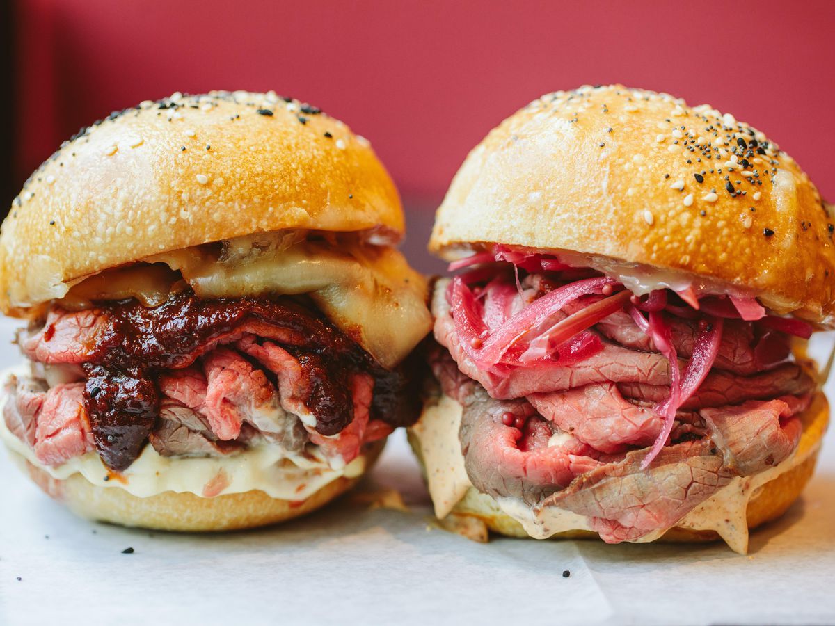 Two roast beef sandwiches sit side by side, showing off thinly slices rare meat and topped with red onions and other condiments