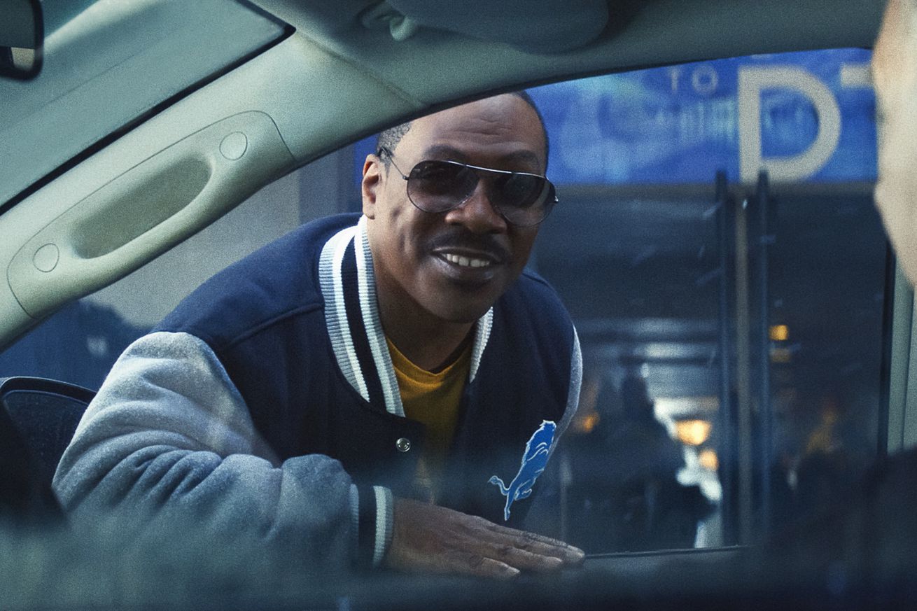 A man wearing aviators and a letterman jacket leaning over to peer into the window of a parked car.