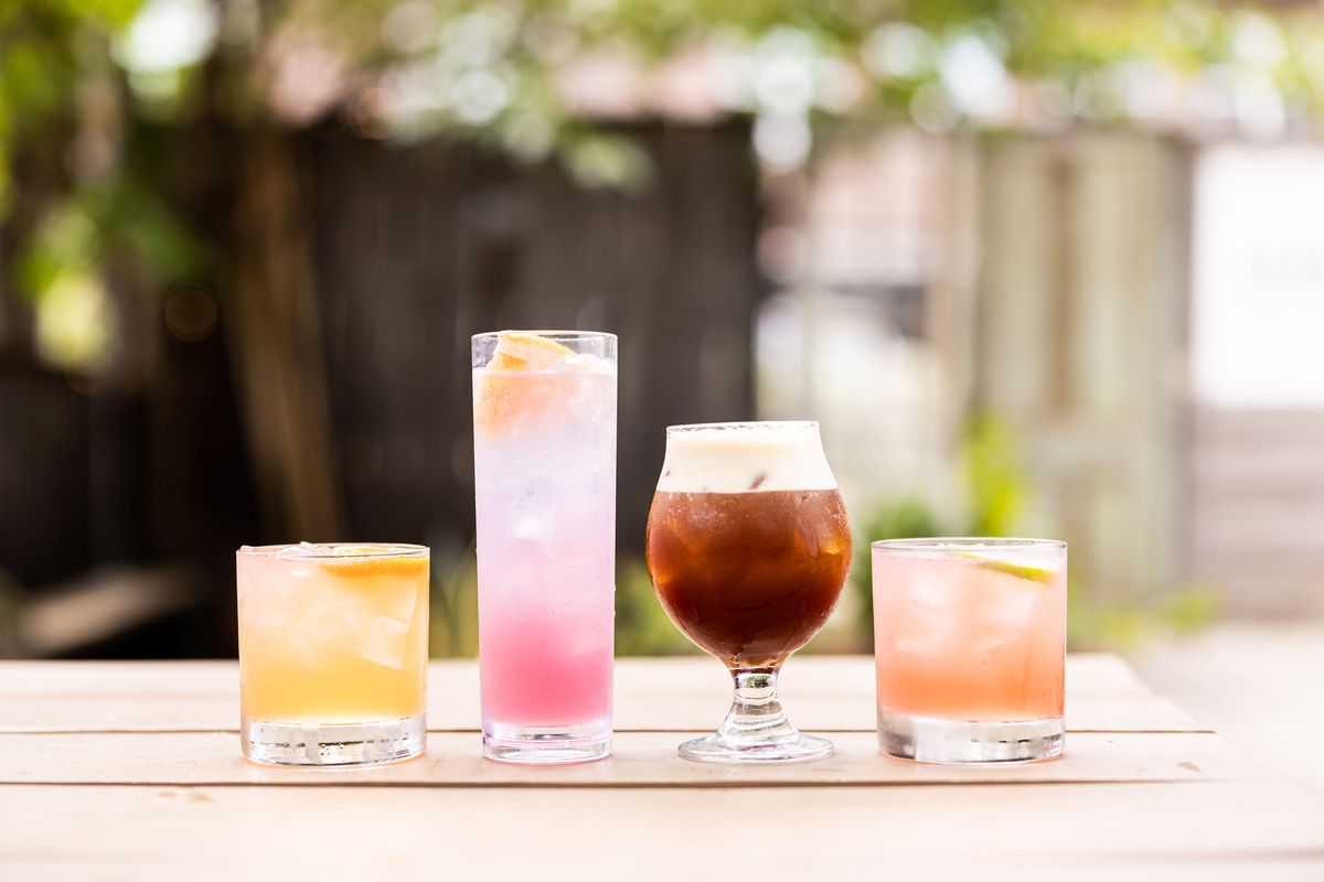 Four cocktails are lined up on a picnic table, in various colors and different types of glasses.
