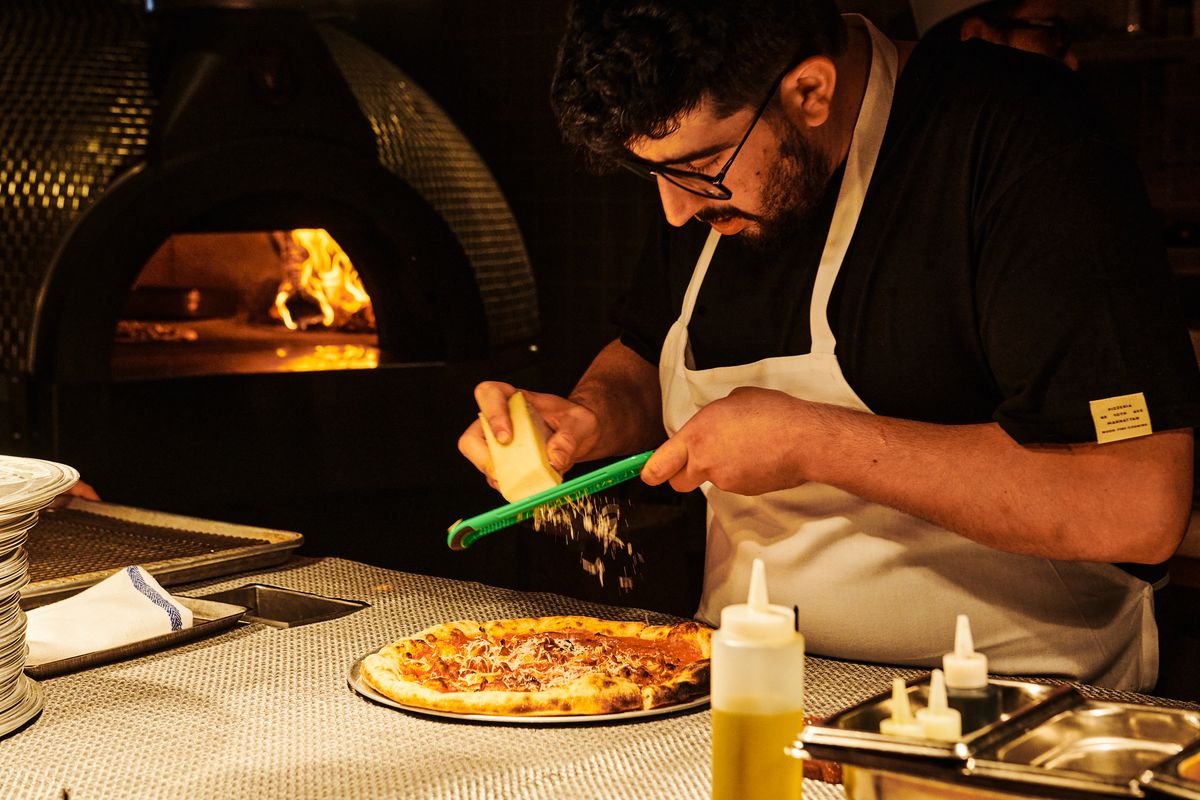 A bespectacled staffer shaves pecorino over a guanciale pie while flames are visible from the domed pizza oven behind him