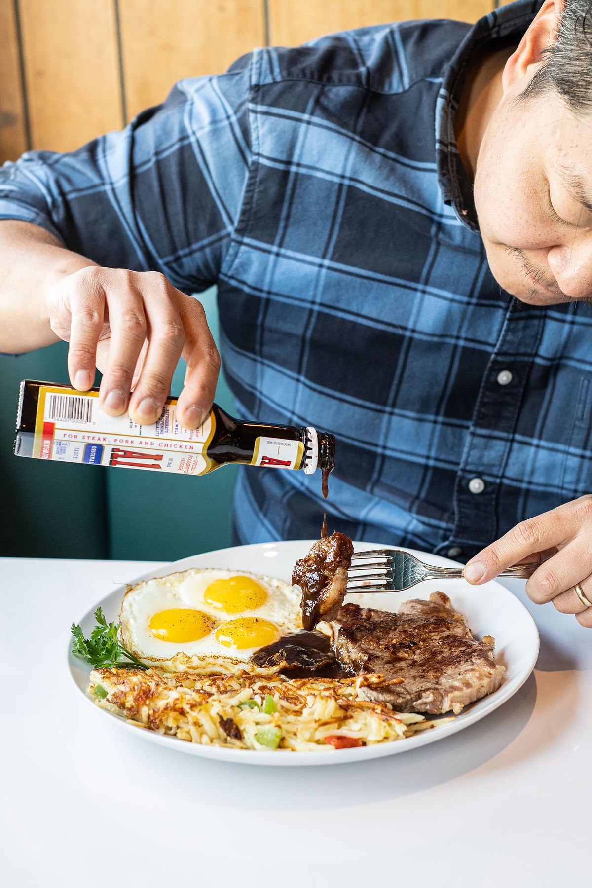Man pouring bottle of A1 sauce on a plate containing eggs, steak, and hash browns.