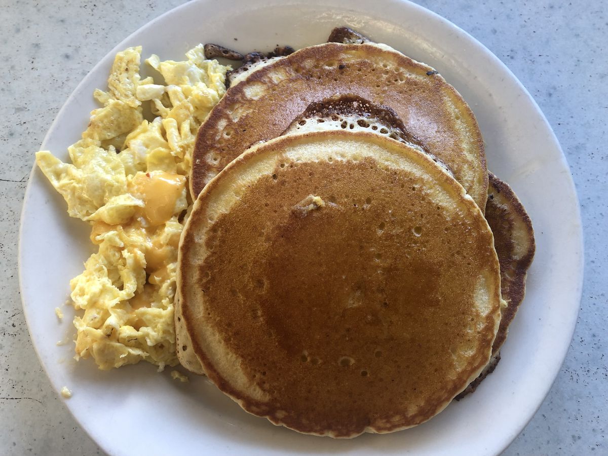 Two large, crispy-edged pancakes and a side of cheesy scrambled eggs on a circular white plate set on top of a white countertop.