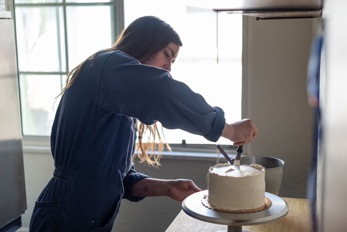 Woman wearing jumpsuit applies white icing to the outside of a cake sitting on a cake stand.