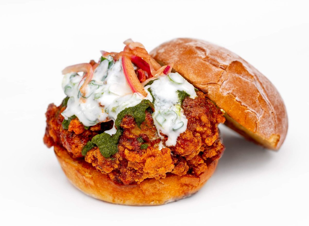 A fried chicken sandwich piled with white and green sauces plus diced onions against a white background.