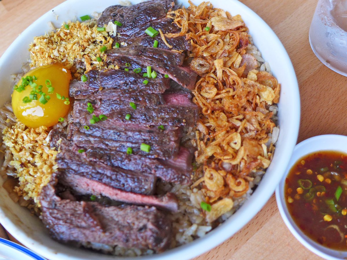 A dish with slices of steak, rice, and fried shallots with a dish of sauce on the side.