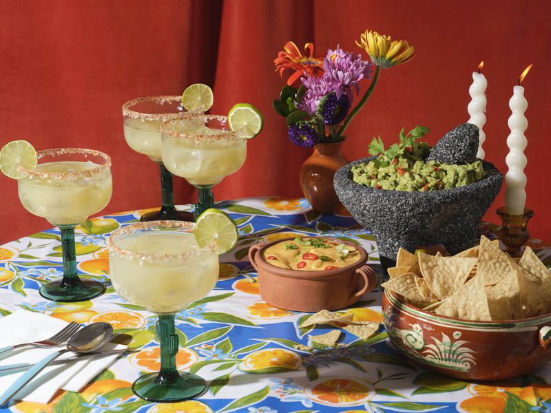 A table set with glasses of margaritas, guacamole in a molcajete, a bowl of tortilla chips, and queso.