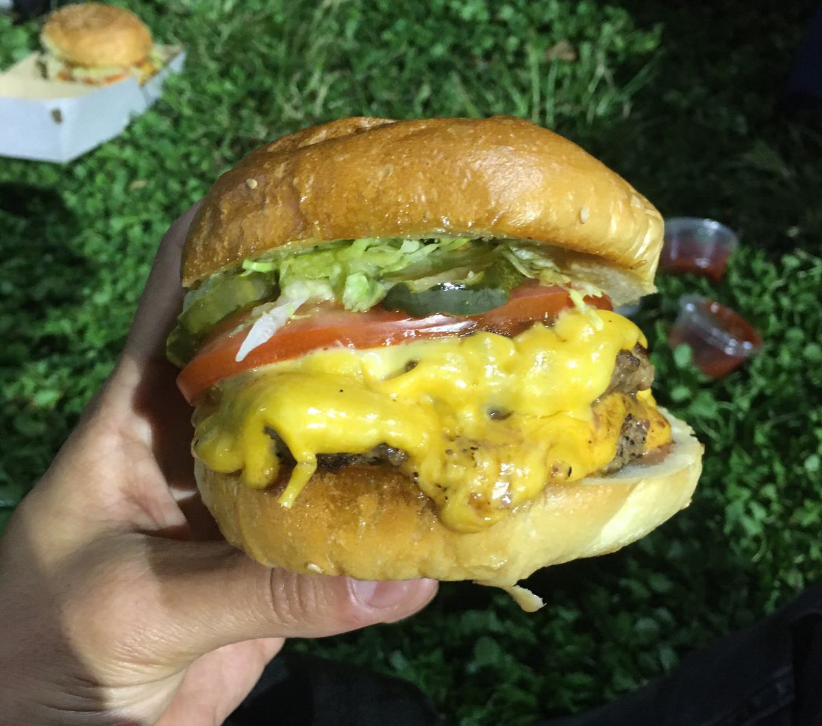 A hand holds a double cheeseburger overflowing with melty American cheese, scraps of lettuce, pickles, and tomato against a background of grass