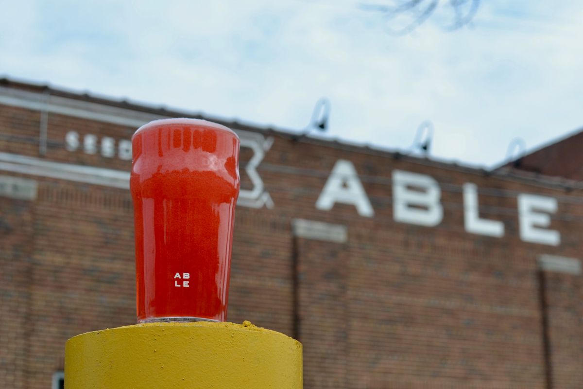 A reddish beer in a pint glass in front of a brick building with the word “Able” painted on it in white paint. 