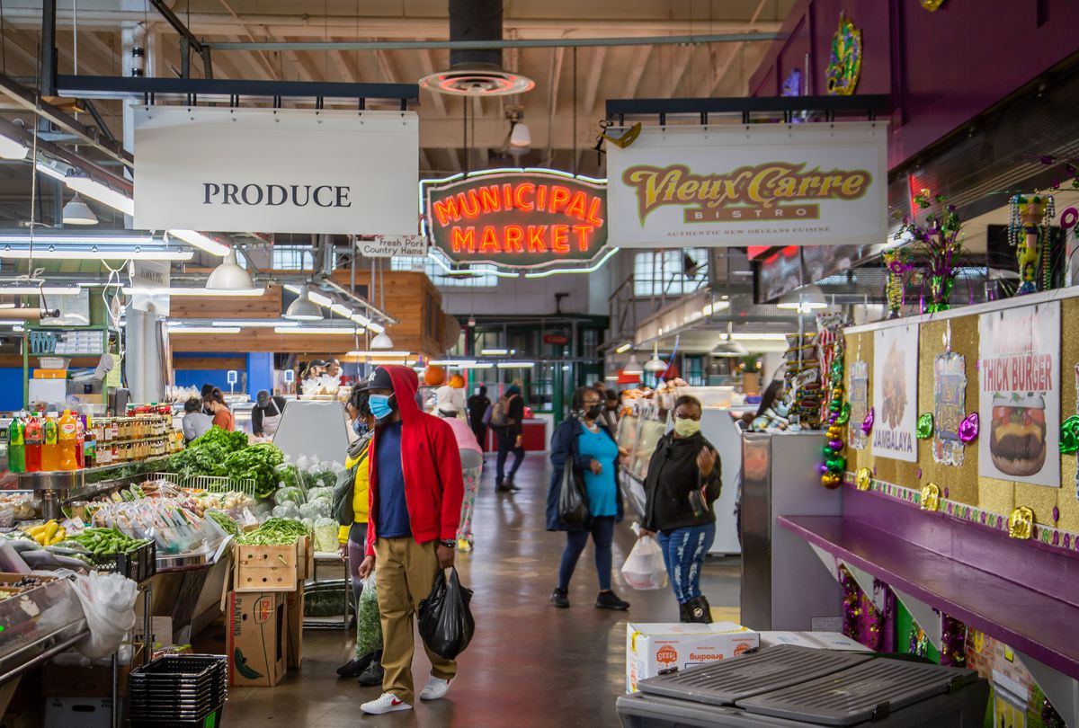 Shoppers perusing the produce and meat stalls inside the Municipal Market of Atlanta, the Sweet Auburn Curb Market.  The old red neon sign “Municipal Market” hangs above the crowd, with signs for “Produce” and New Orleans food stall “Vieux Carre Bistro” hanging in the foreground. 