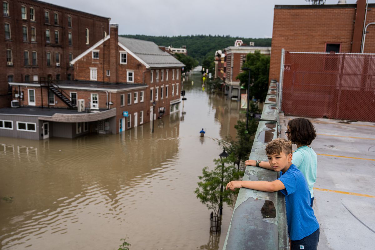 Two children stand on the roof of a building in Montpelier, Vermont. The streets below are flooded with brown water.