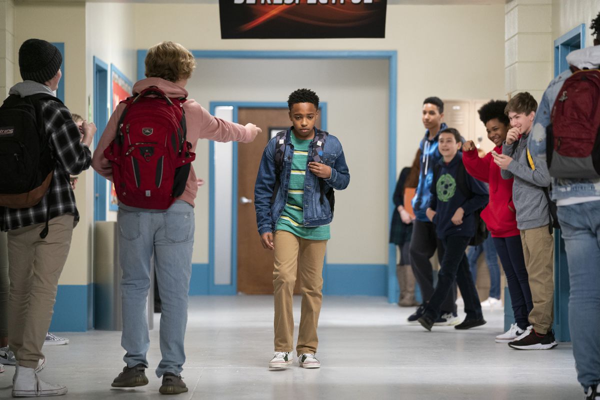 A junior-high age boy with a backpack over one shoulder walks alone in the hall as someone points at him