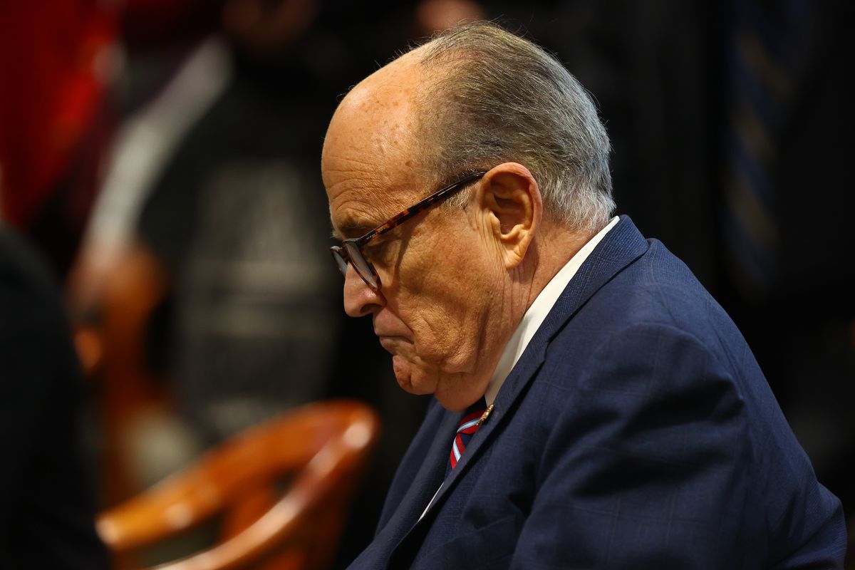 Giuliani, in tortoise shell glasses, a navy suit, white shirt, and red and blue tie, sits with his chin on his chest. He appears to be frowning slightly, as if deep in thought. A number of blurred figures are behind him.