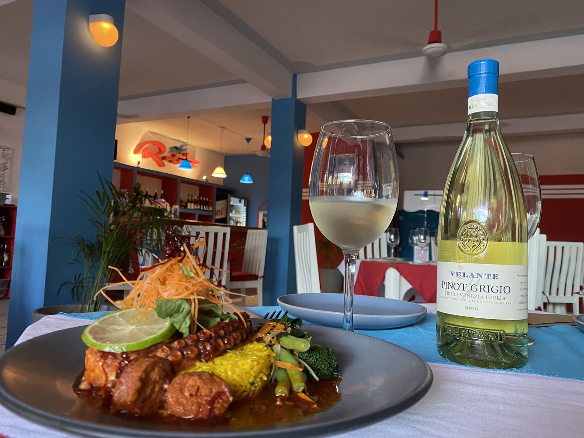 A plate of octopus with lots of fixings in a pool of sauce, beside a glass and bottle of white wine, in a jazzy restaurant interior