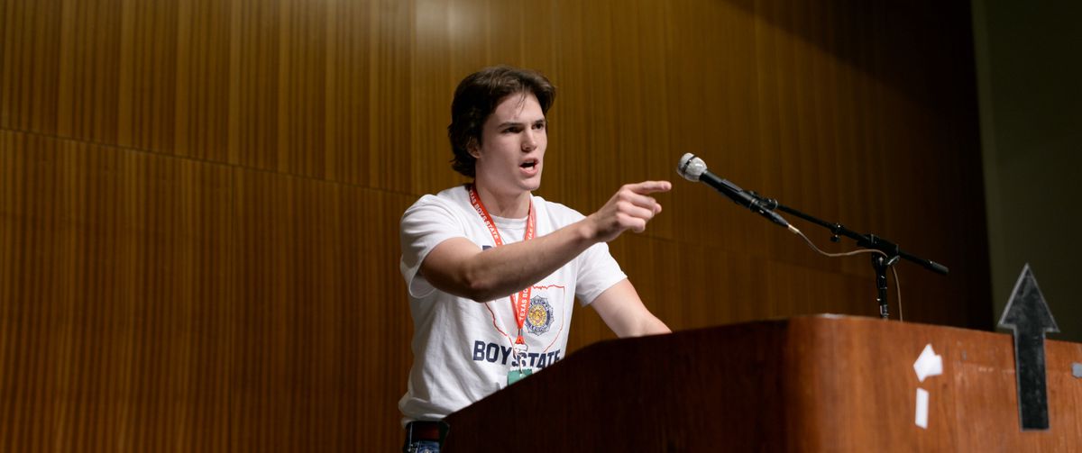 A teenage boy stands at a lectern pointing as he delivers a speech in Boys State