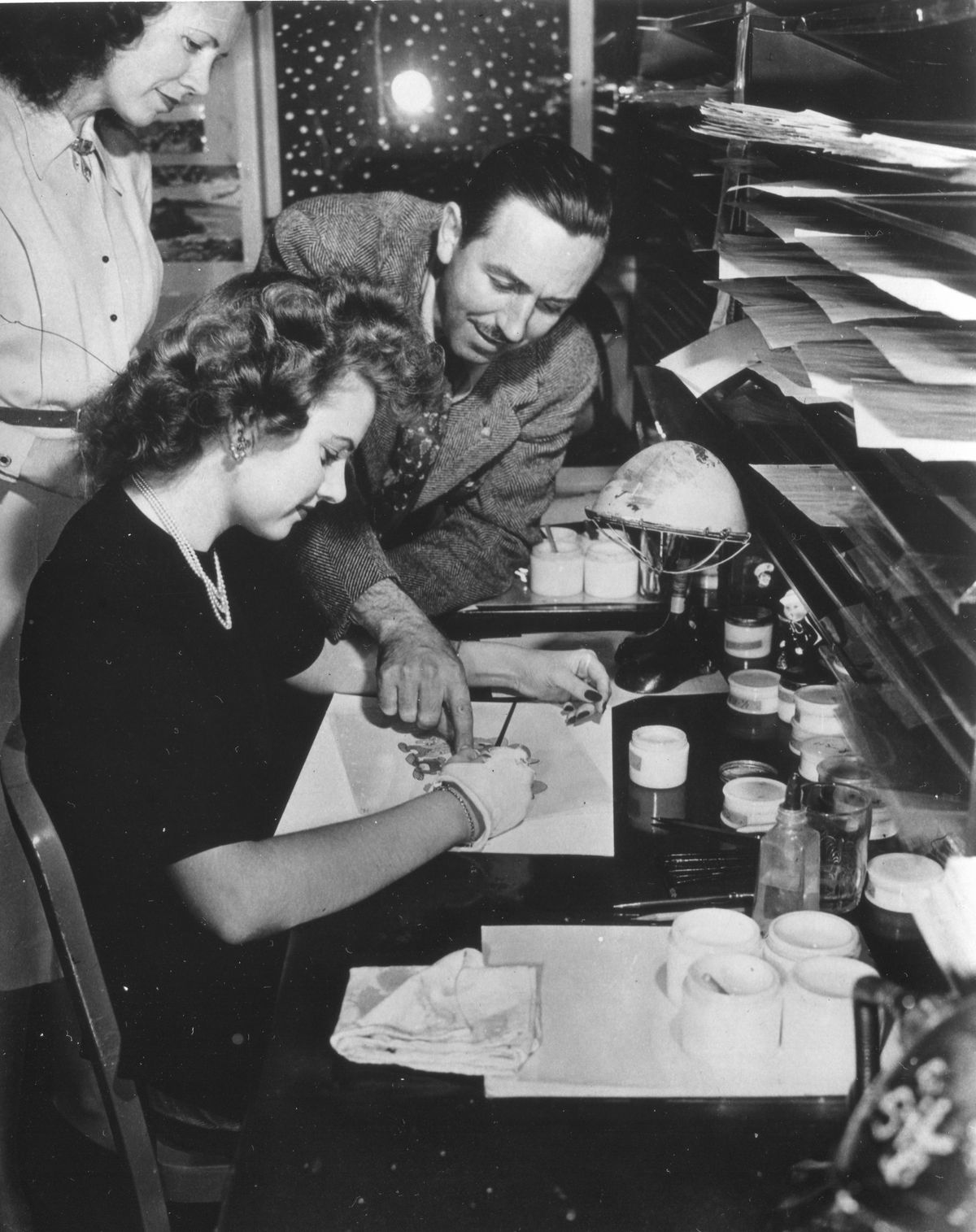 Walt Disney leans over painter Edith Moore as she works at her desk on an animation cel from an unspecified film in an archival photo from 1943