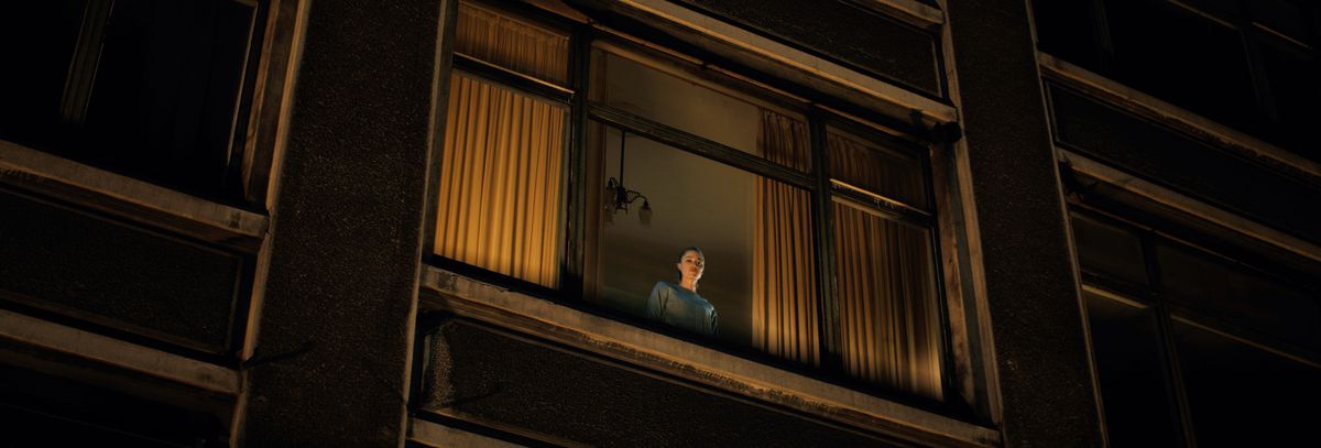 Maika Monroe stands as a tiny figure looking out of a high window at night in Watcher