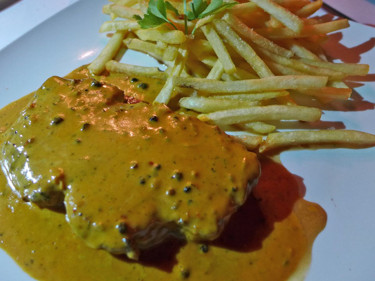 A slab of steak doused in a mustard color sauce with a side of french fries.