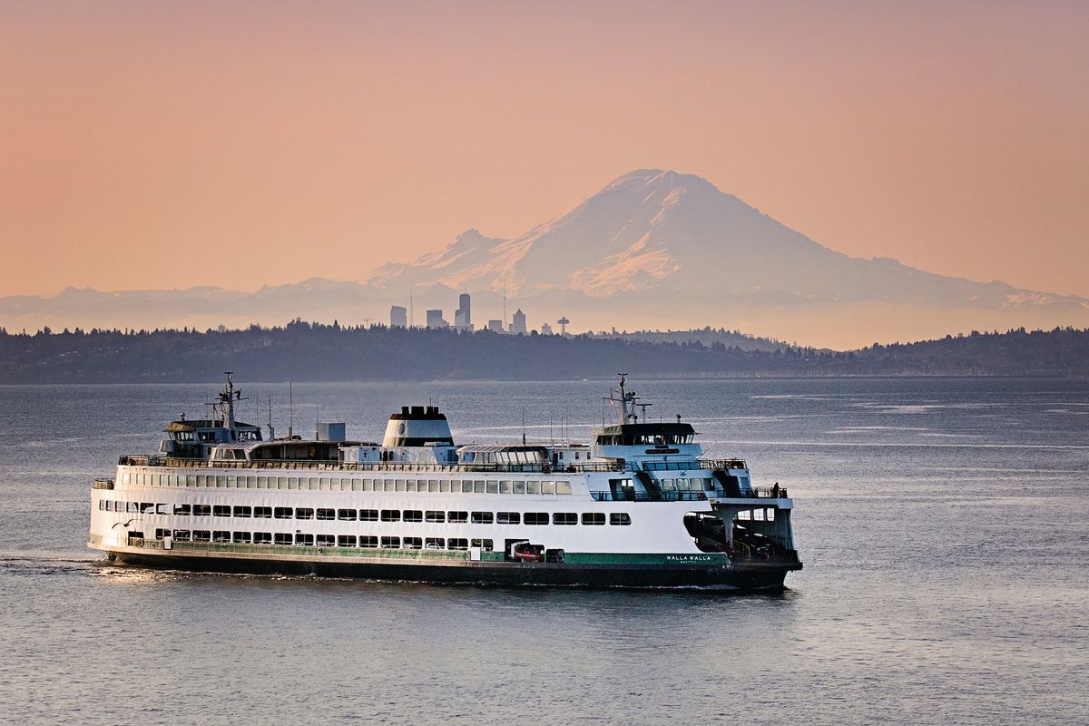 A large white ferry with green trim on calm waters. There’s a large mountain in the background and silhouettes of hills and skyscrapers.