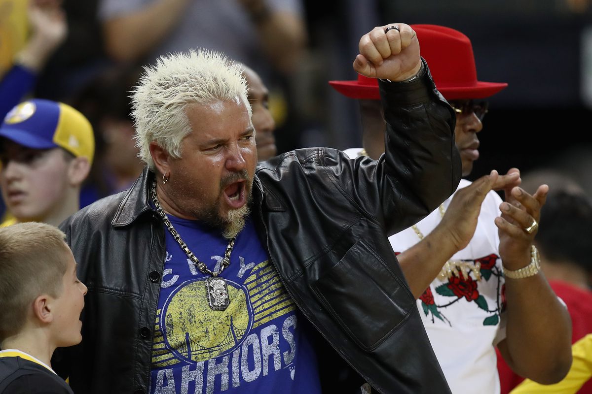 Guy Fieri in a Golden State Warriors t-shirt, gold chain, and black leather jacket, pumps his fist while sitting courtside at an NBA basketball game.