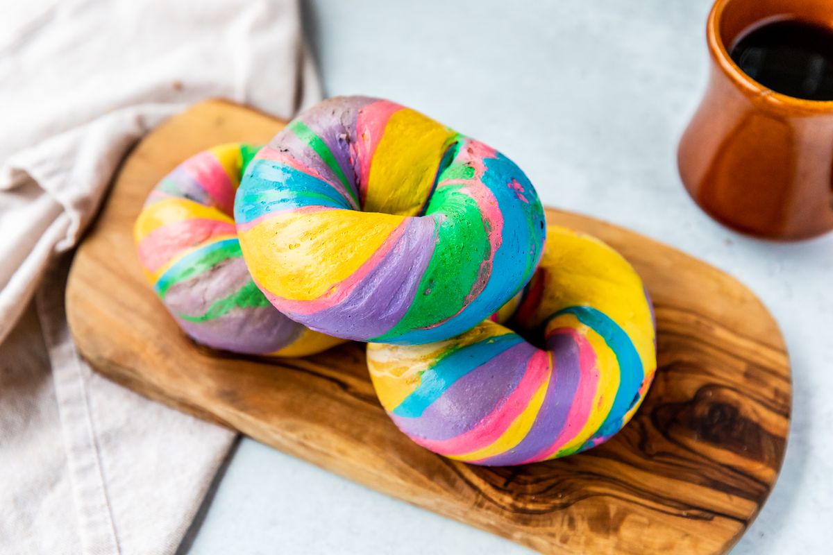 Plain bagels with multicolor swirls sitting on a board next to a cup of coffee.