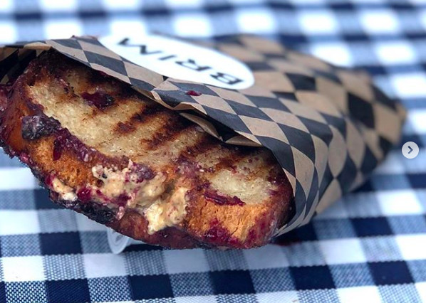 A crusted, toasted sandwich, wrapped in paper with a Brim sticker on a blue and white gingham tablecloth. Inside the sandwich gooey nut butter and purple blue berry jam oozes out.