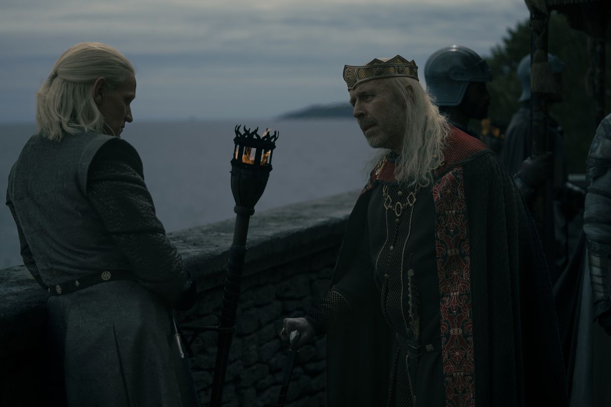 Daemon leaning against a wall with his back slightly to the camera while Viserys stands in front of him, leaning on a cane and looking consternated