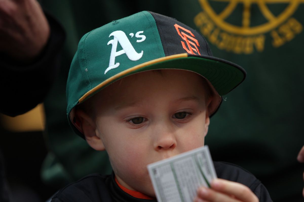 Charlie Chesnosky, 3, of Burllingame, studies a baseball card while wearing his split bill A’s-Giants hat before the first game of the Bay Bridge spring training series between the Oakland Athletics and the San Francisco Giants Thursday March 27, 2014, at