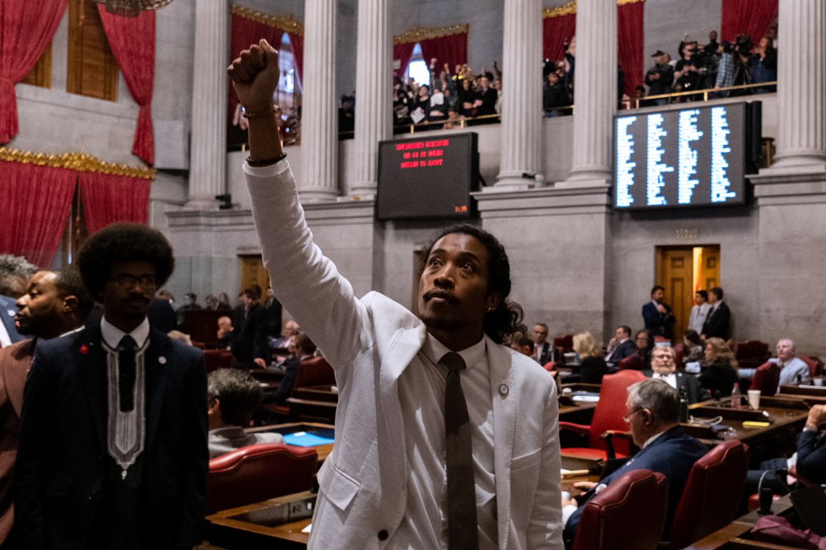 Rep. Justin Jones, a Black man in a light gray suit standing on the floor of the state legislature, raises a fist in recognition to supporters above him in the gallery.