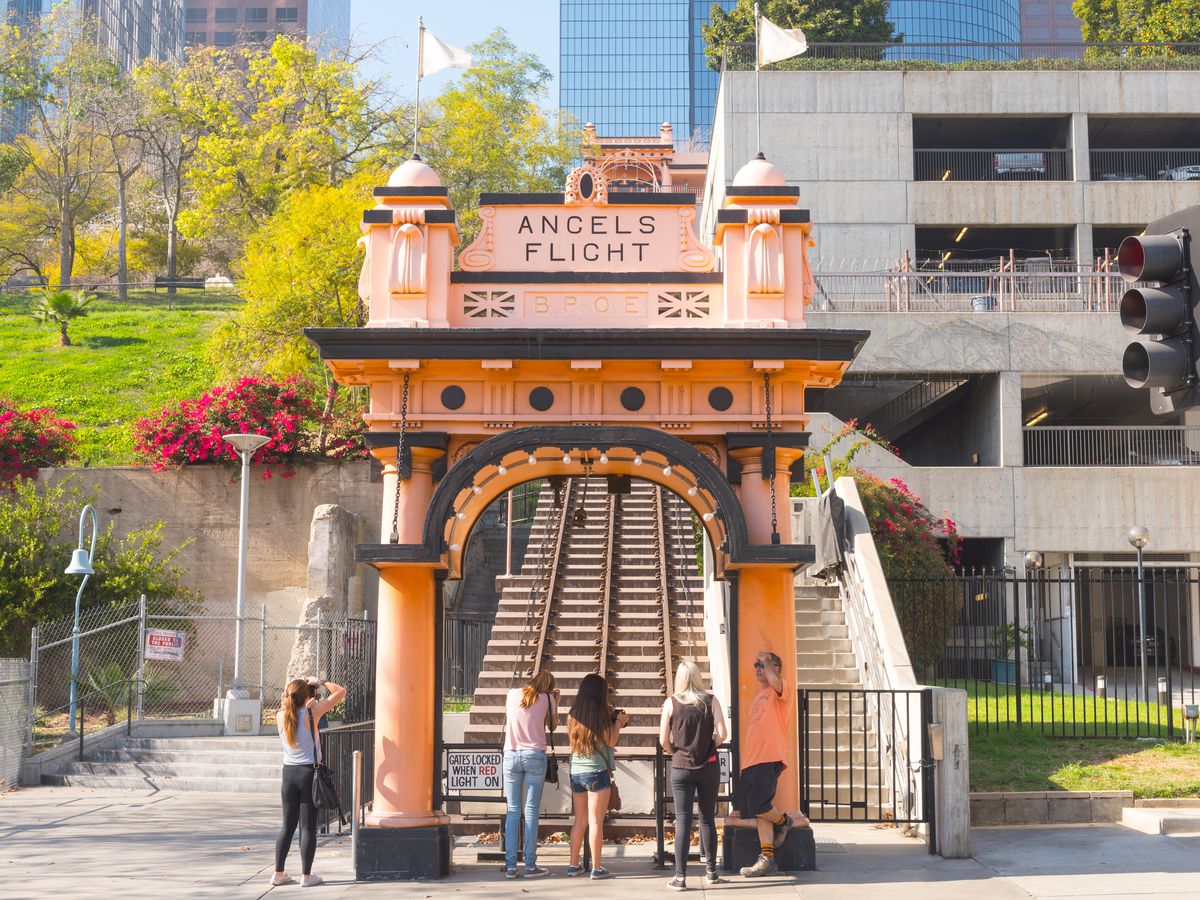 In the foreground is the entrance to a railway with train tracks. The sign on the entrance reads Angels Flight. Behind the railway are buildings and a park with trees and flowers.