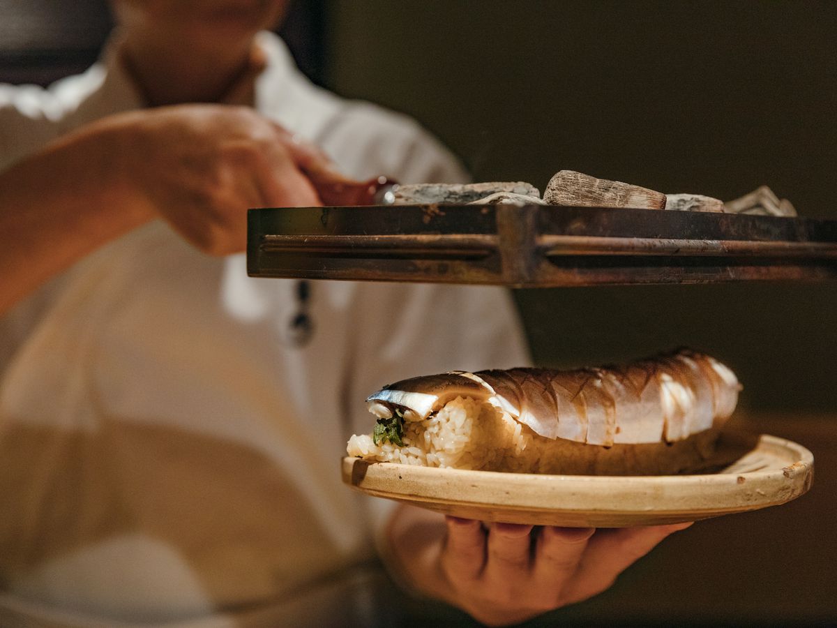 A chef in white uniform holds a hand-held grill containing binchotan charcoal over a plate of mackeral.