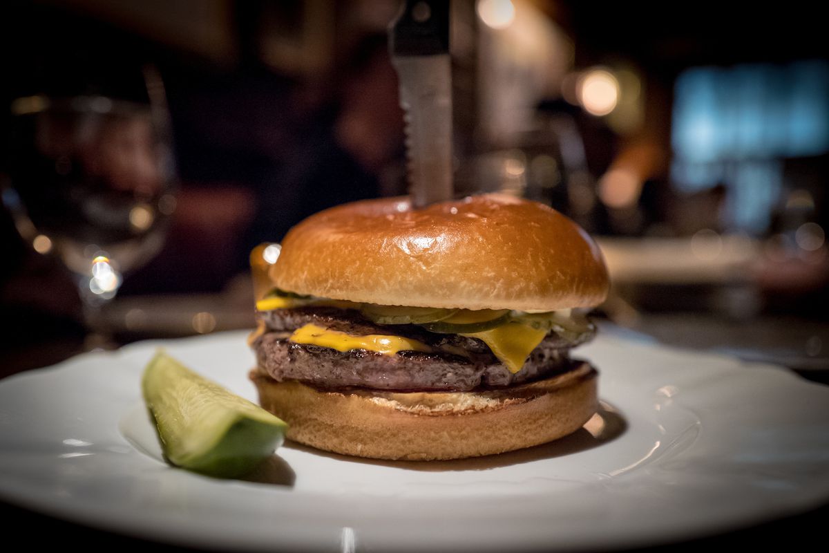 A knife impales a burger, which features two thin, griddled, four-ounce patties, Kraft American cheese, Dijonnaise, and pickles