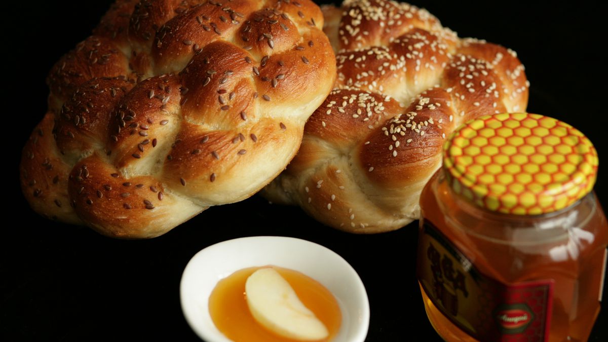 Two round braided challah next to a bowl of honey with a single apple slice in it and a jar of honey, on a black background. 
