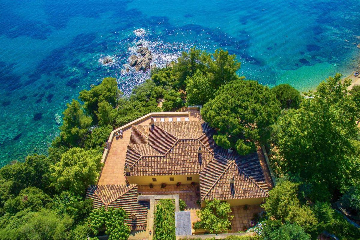 Aerial view of terra cotta-roofed villa perched on a hill above a dazzling blue sea surrounded by trees.