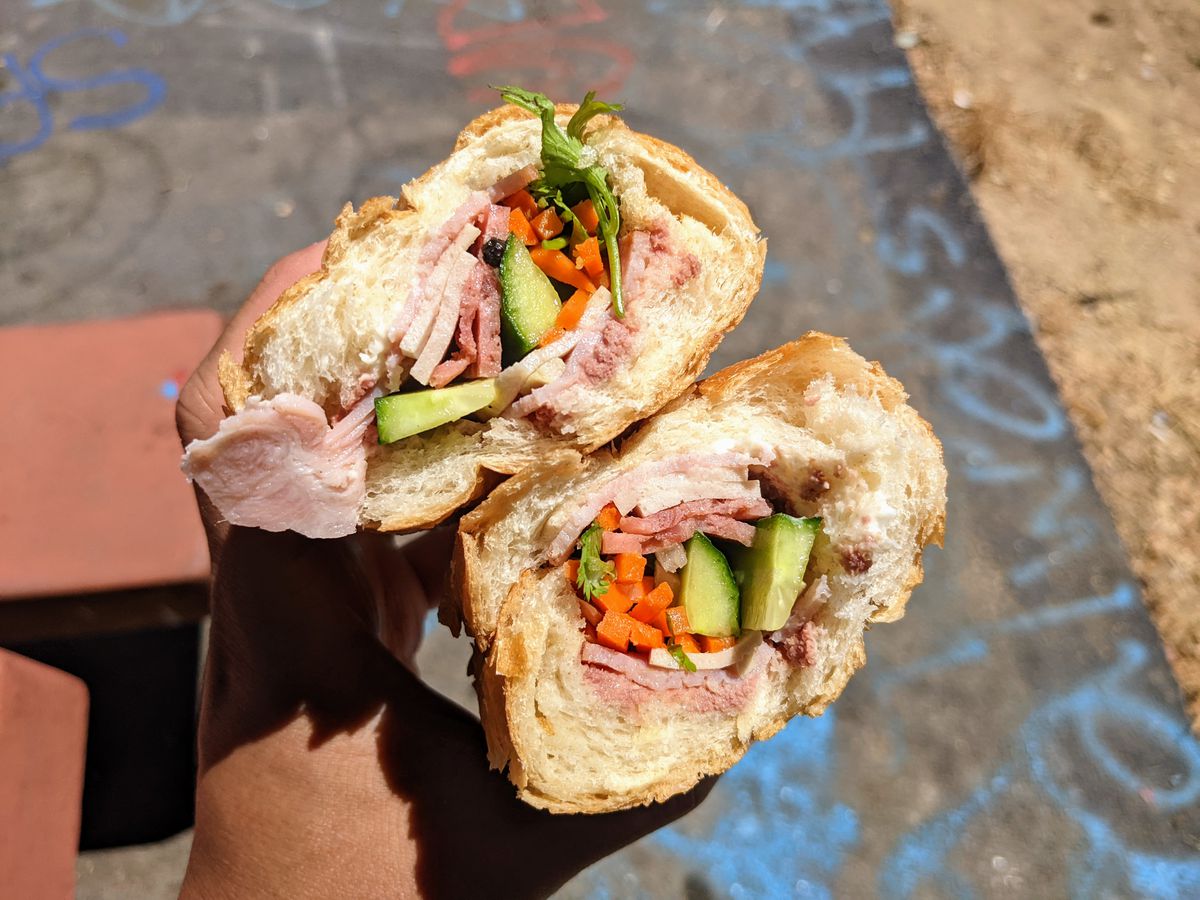 Split banh mi held in hand with sliced meats and pickled vegetables.
