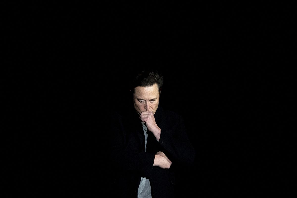 Elon Musk standing on a dark stage with his chin in his hand.