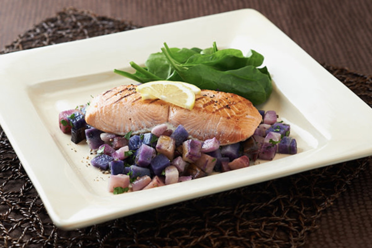 A cooked salmon on a plate with a lemon slice on top and roasted purple-colored potatoes on the side
